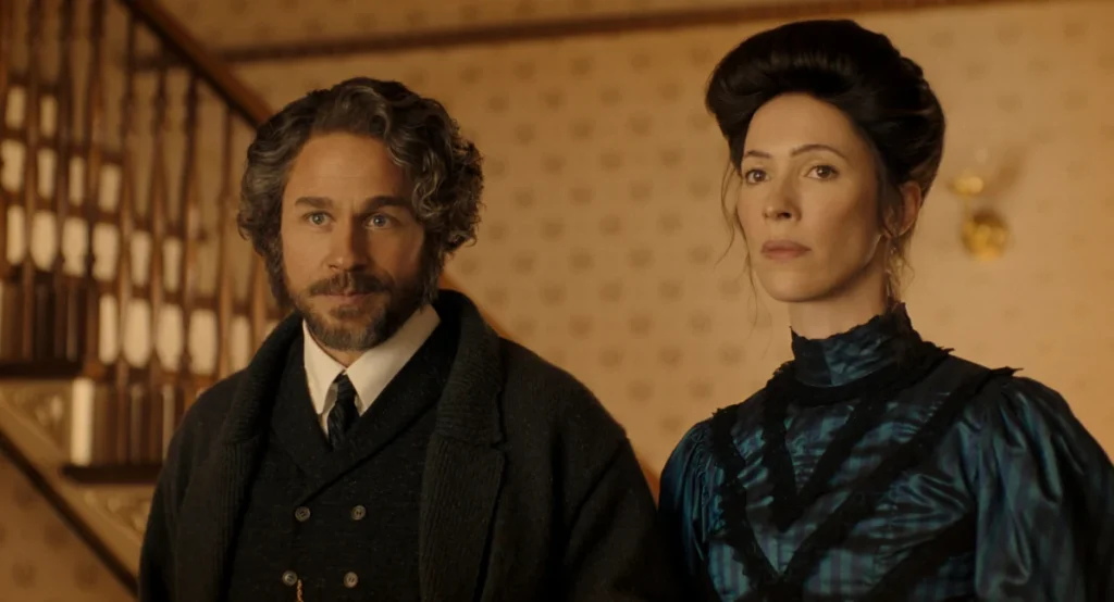 "A man with curly hair and a beard stands beside a woman with dark hair in an old-fashioned, elegant dress, both in a warmly lit vintage interior with patterned wallpaper and a staircase in the background."