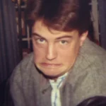 "Young man indoors making a humorous, exaggerated face, wearing a grey jacket with leather sleeves. The environment looks like a living room with curtains and patterned furniture in the background. Warm, casual setting."